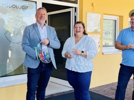 Martina gemeinsam mit dem Bürgermeister bei der Wappenübergabe Drei Personen stehen vor einem Gebäude und lächeln, einer hält einen bunten Korb.