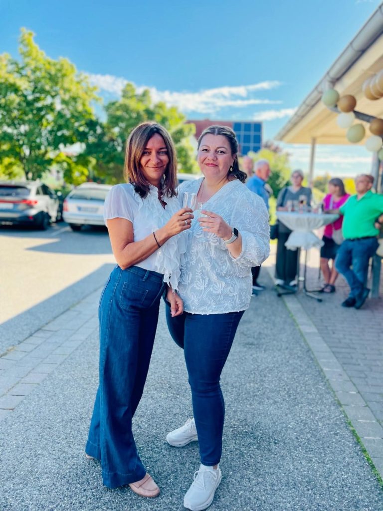 Martina und Sandra Gerbholz von SINN & MARKE in guter Stimmung Zwei Frauen stehen neben einer Gruppe von Menschen, sonniger Tag, entspannte Atmosphäre.